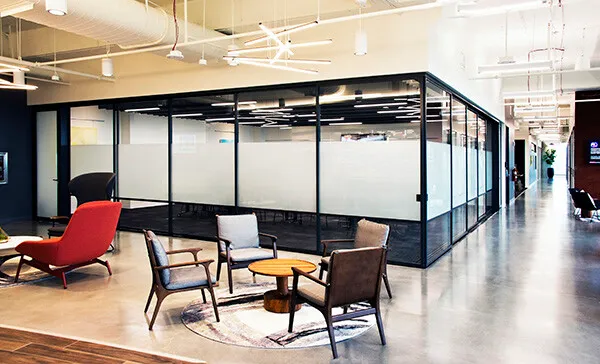 Modern office space with glass-walled meeting room, several chairs around small tables, and overhead lighting fixtures. Carpeted area contrasts with polished concrete floor.