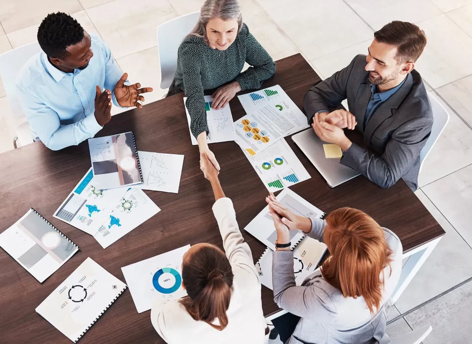 A diverse group of Five people sit around a table in a meeting, discussing documents and charts.